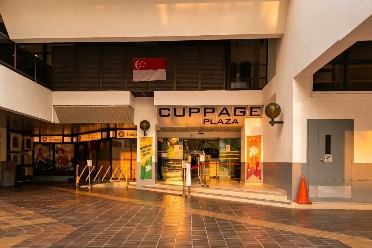 The image captures the entrance of Cuppage Plaza in Singapore under warm evening light, featuring a Singapore flag hanging above the entryway. A restaurant named Shinjuku is visible on the left, while the tiled foreground reflects the golden sunlight. The image captures the entrance of Cuppage Plaza in Singapore under warm evening light, featuring a Singapore flag hanging above the entryway. A restaurant named Shinjuku is visible on the left, while the tiled foreground reflects the golden sunlight.