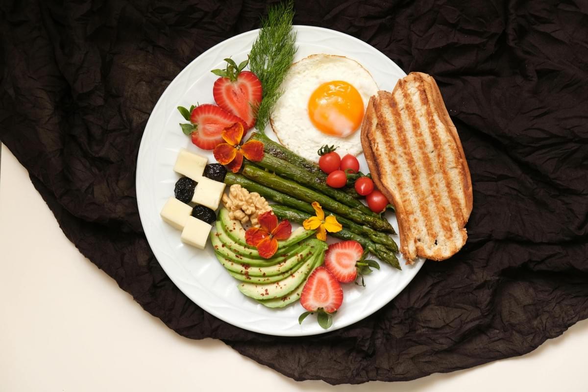 A white plate filled with a colorful breakfast features a sunny-side-up egg, grilled bread, roasted asparagus spears, sliced avocado, and fresh strawberries. The plate is also garnished with cherry tomatoes, cheese cubes, walnuts, and edible flowers, all set against a dark, textured cloth background. A white plate filled with a colorful breakfast features a sunny-side-up egg, grilled bread, roasted asparagus spears, sliced avocado, and fresh strawberries. The plate is also garnished with cherry tomatoes, cheese cubes, walnuts, and edible flowers, all set against a dark, textured cloth background.