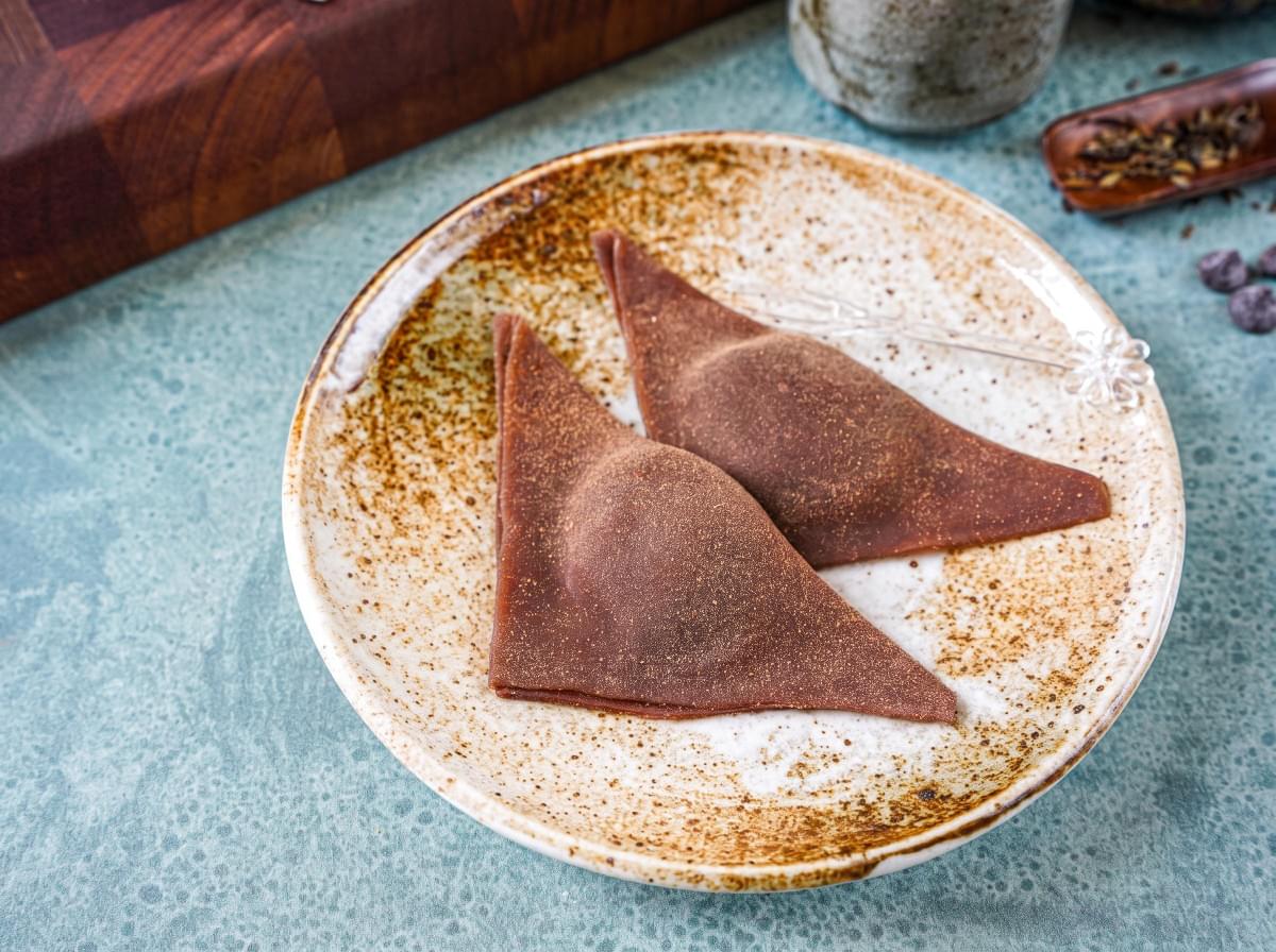 Two cinnamon-dusted triangular yatsuhashi pieces on a ceramic plate with a small pick. Two cinnamon-dusted triangular yatsuhashi pieces on a ceramic plate with a small pick.