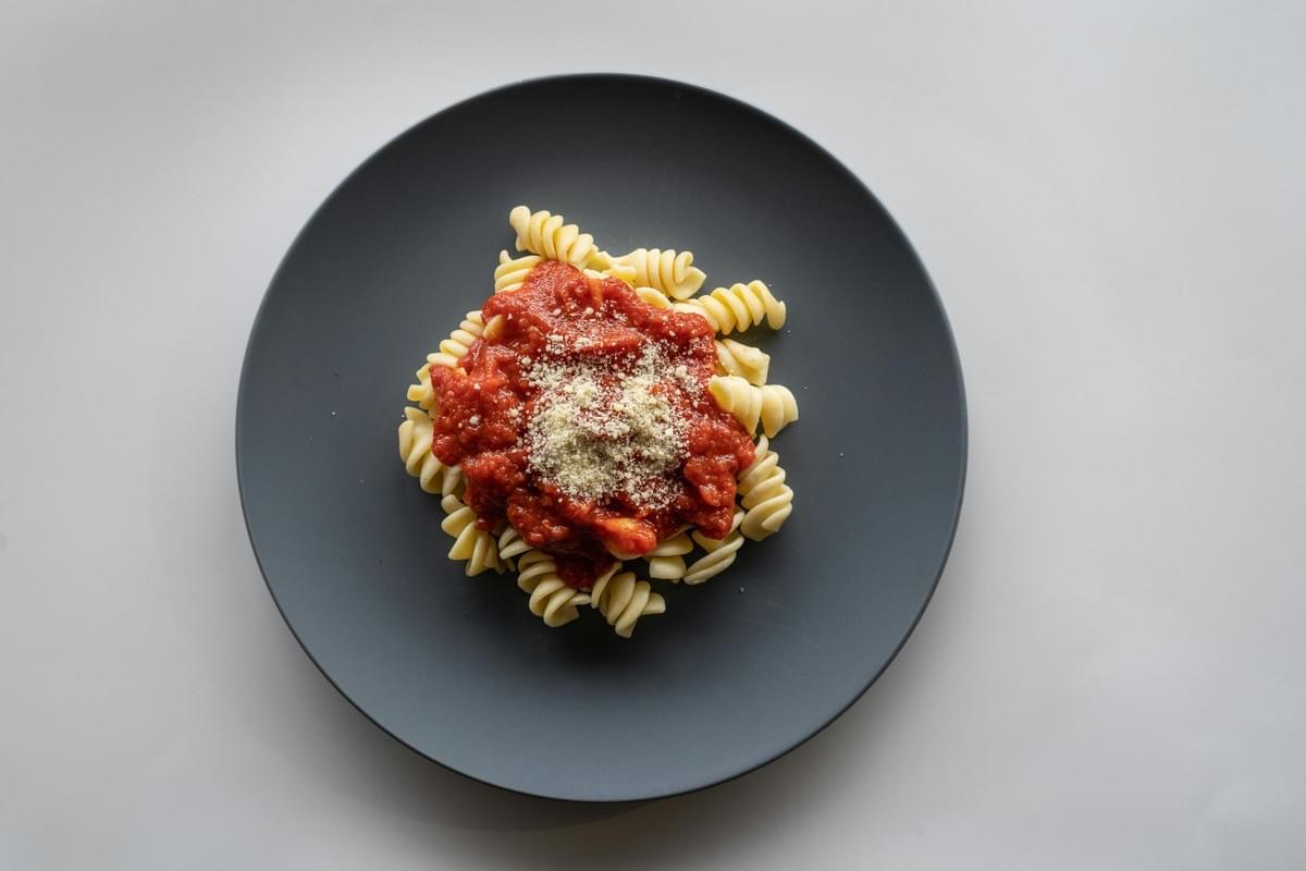 A top-down view shows a serving of spiral rotini pasta centered on a dark matte grey plate against a light background. Thick, textured red marinara sauce covers the middle of the pasta and is topped with a dusting of grated parmesan cheese. A top-down view shows a serving of spiral rotini pasta centered on a dark matte grey plate against a light background. Thick, textured red marinara sauce covers the middle of the pasta and is topped with a dusting of grated parmesan cheese.