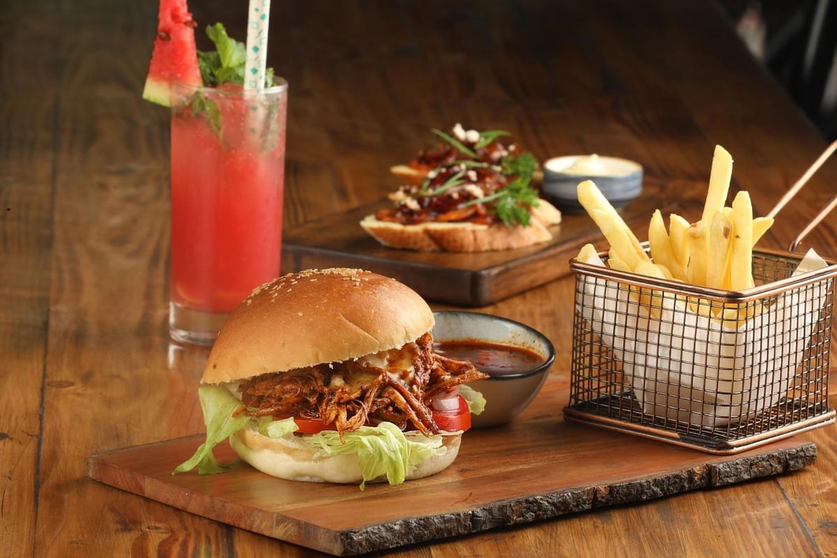 A pulled meat burger with lettuce and tomato is served on a wooden board alongside a wire basket of french fries and a small bowl of dipping sauce. In the background, a vibrant red watermelon drink with a mint garnish sits near a plate of bruschetta toppings. A pulled meat burger with lettuce and tomato is served on a wooden board alongside a wire basket of french fries and a small bowl of dipping sauce. In the background, a vibrant red watermelon drink with a mint garnish sits near a plate of bruschetta toppings.