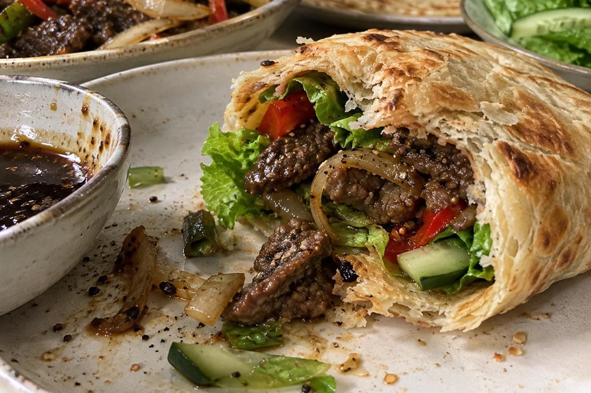 A macro, close-up view of a partially eaten black pepper beef roti wrap on a ceramic plate, highlighting its flaky, layered pastry-like texture. The cross-section reveals tender stir-fried beef coated in a dark, peppered sauce, combined with crisp green lettuce, sautéed onions, red bell pepper strips, and cucumber slices. Next to the wrap, a small dipping bowl contains dark black pepper sauce with floating specks of cracked pepper and green onion. A macro, close-up view of a partially eaten black pepper beef roti wrap on a ceramic plate, highlighting its flaky, layered pastry-like texture. The cross-section reveals tender stir-fried beef coated in a dark, peppered sauce, combined with crisp green lettuce, sautéed onions, red bell pepper strips, and cucumber slices. Next to the wrap, a small dipping bowl contains dark black pepper sauce with floating specks of cracked pepper and green onion.