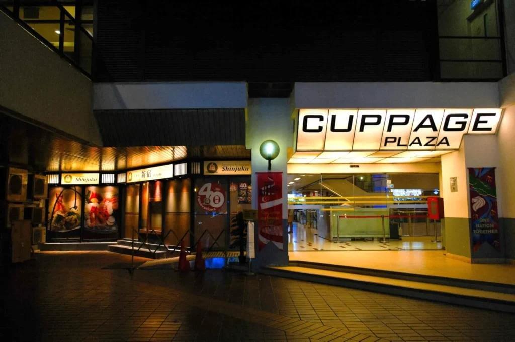An evening view of the brightly lit entrance to Cuppage Plaza, featuring a prominent illuminated sign with the building's name above glass doors. To the left, a Japanese restaurant named Shinjuku displays glowing food imagery alongside signs celebrating its 40th anniversary. An evening view of the brightly lit entrance to Cuppage Plaza, featuring a prominent illuminated sign with the building's name above glass doors. To the left, a Japanese restaurant named Shinjuku displays glowing food imagery alongside signs celebrating its 40th anniversary.