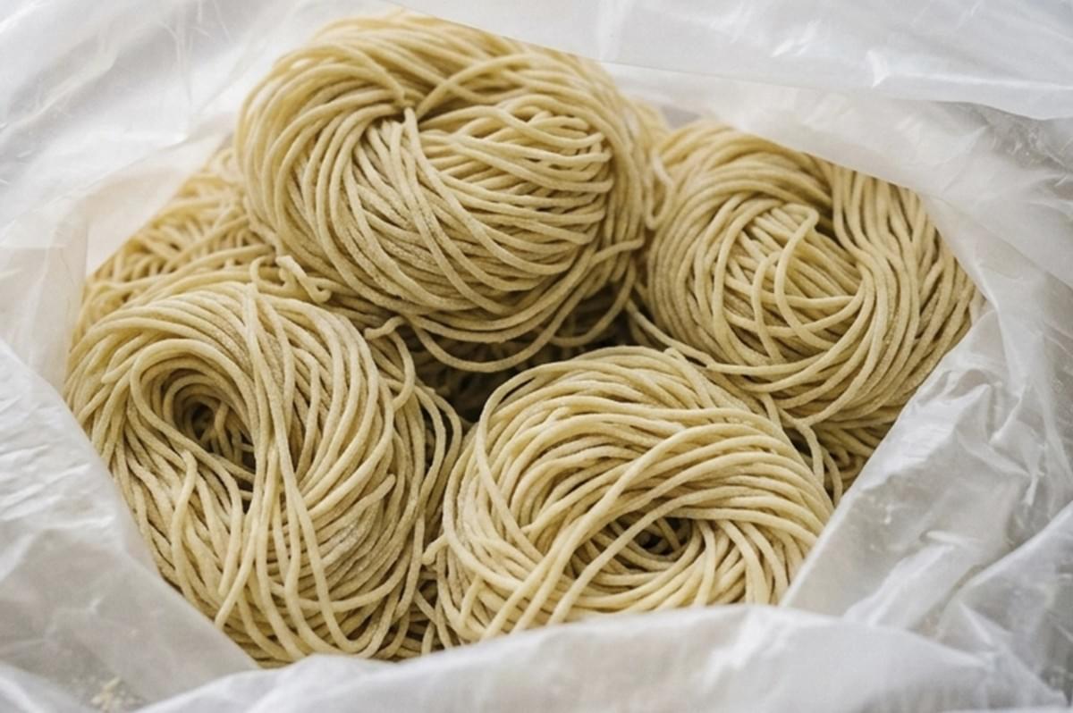 A top-down close-up of several bundles of fresh, uncooked ramen noodles. The thin, pale-yellow noodles are neatly swirled into circular nests and stored inside a clear plastic bag. The texture of the flour-dusted strands is highly visible, emphasizing a "behind-the-scenes" culinary preparation vibe with soft, even lighting. A top-down close-up of several bundles of fresh, uncooked ramen noodles. The thin, pale-yellow noodles are neatly swirled into circular nests and stored inside a clear plastic bag. The texture of the flour-dusted strands is highly visible, emphasizing a "behind-the-scenes" culinary preparation vibe with soft, even lighting.