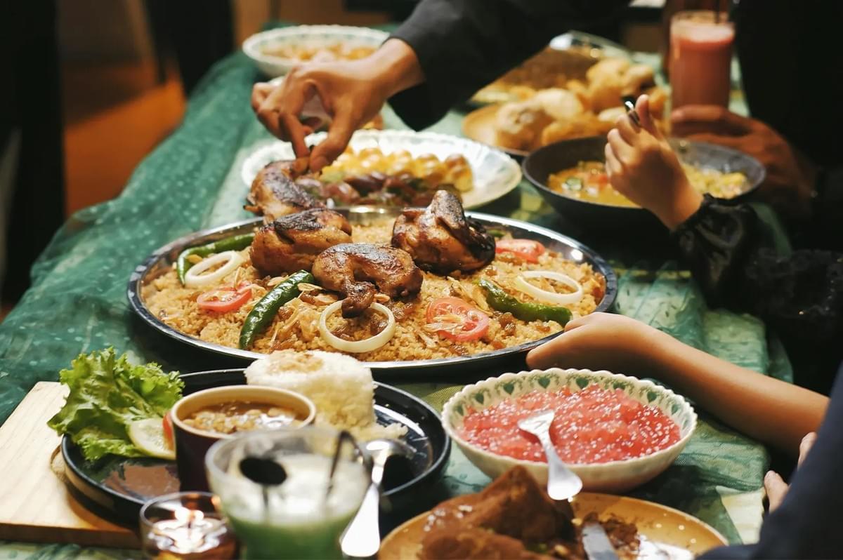 A communal dinner feast, featuring a large platter of grilled chicken over mandhi rice with tomatoes and onions, surrounded by various side dishes and drinks. A communal dinner feast, featuring a large platter of grilled chicken over mandhi rice with tomatoes and onions, surrounded by various side dishes and drinks.