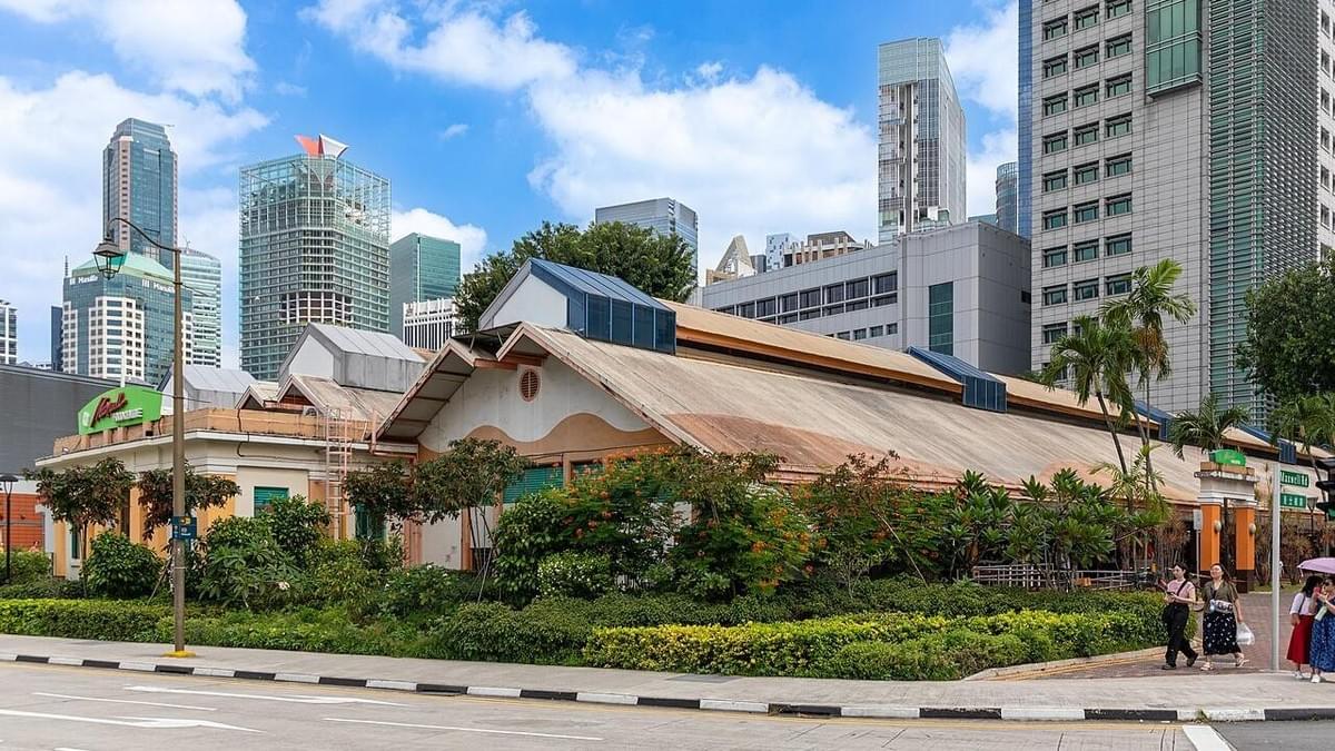 This image shows the exterior of the Maxwell Food Centre in Singapore, featuring its distinctive long, low-slung roof and surrounding greenery set against a backdrop of modern city skyscrapers. This image shows the exterior of the Maxwell Food Centre in Singapore, featuring its distinctive long, low-slung roof and surrounding greenery set against a backdrop of modern city skyscrapers.