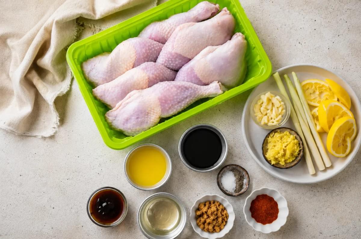 "A green tray of raw chicken legs surrounded by small bowls of marinade ingredients, lemon slices, and lemongrass stalks.” "A green tray of raw chicken legs surrounded by small bowls of marinade ingredients, lemon slices, and lemongrass stalks.”