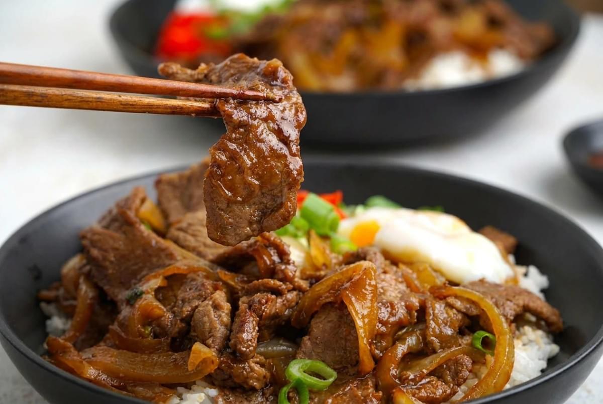 Chopsticks holding up a slice of glazed beef over a rice bowl. Chopsticks holding up a slice of glazed beef over a rice bowl.