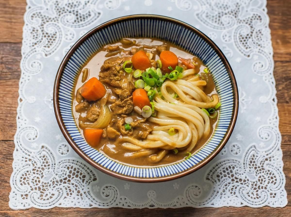 Bowl of curry udon with sliced meat and carrots served on a white lace placement over a wooden table. Bowl of curry udon with sliced meat and carrots served on a white lace placement over a wooden table.