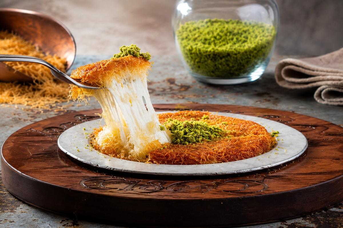 A fork lifting a piece of warm künefe dessert topped with crushed pistachios, stretching gooey melted cheese from the crispy, golden shredded pastry base on a serving plate. A fork lifting a piece of warm künefe dessert topped with crushed pistachios, stretching gooey melted cheese from the crispy, golden shredded pastry base on a serving plate.