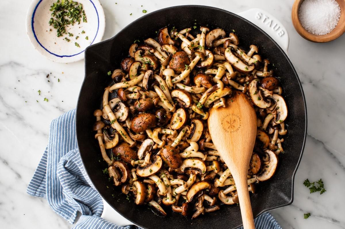 Sautéed mixed mushrooms garnished with chopped herbs in a cast-iron skillet with a wooden spoon. Sautéed mixed mushrooms garnished with chopped herbs in a cast-iron skillet with a wooden spoon.