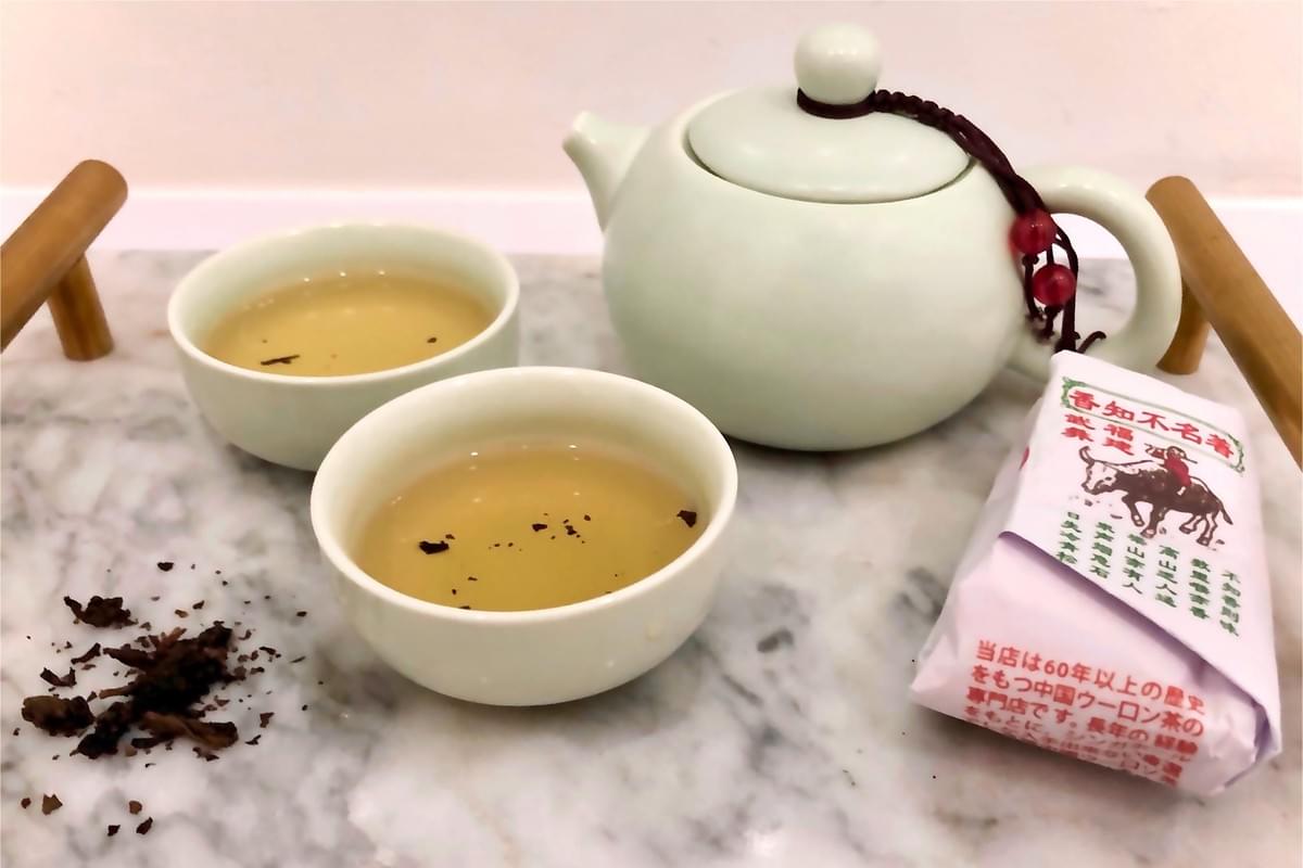 A light green ceramic teapot and two matching cups of tea sit on a marble tray with handles. Loose tea leaves are scattered on the surface next to a small white packet of tea. A light green ceramic teapot and two matching cups of tea sit on a marble tray with handles. Loose tea leaves are scattered on the surface next to a small white packet of tea.