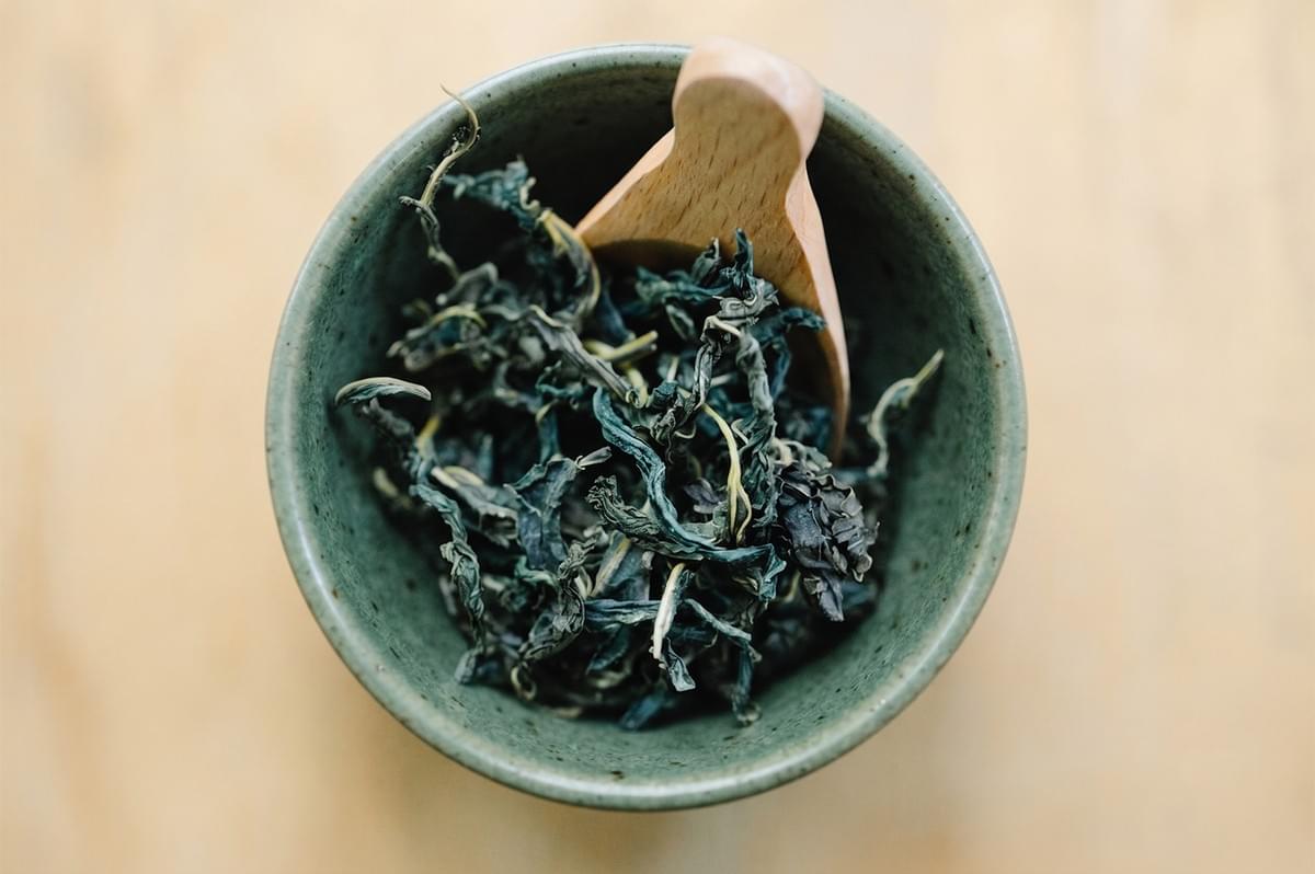 Top-down view of loose-leaf green tea in a rustic ceramic bowl with a small wooden scoop. Top-down view of loose-leaf green tea in a rustic ceramic bowl with a small wooden scoop.
