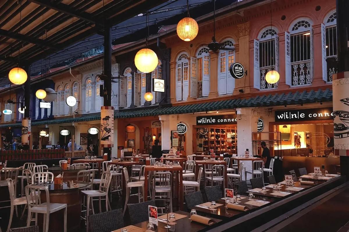 An open-air dining terrace is filled with dining tables illuminated by warm, hanging globe lanterns. In the background, colourful heritage shophouses feature lit storefronts and traditional shuttered windows. An open-air dining terrace is filled with dining tables illuminated by warm, hanging globe lanterns. In the background, colourful heritage shophouses feature lit storefronts and traditional shuttered windows.