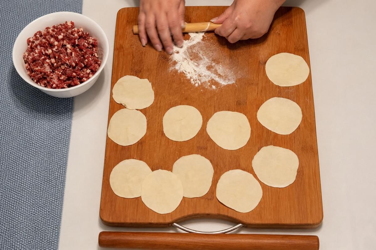 Hands rolling out dough circles on a wooden board next to a bowl of ground meat filling for dumplings Hands rolling out dough circles on a wooden board next to a bowl of ground meat filling for dumplings