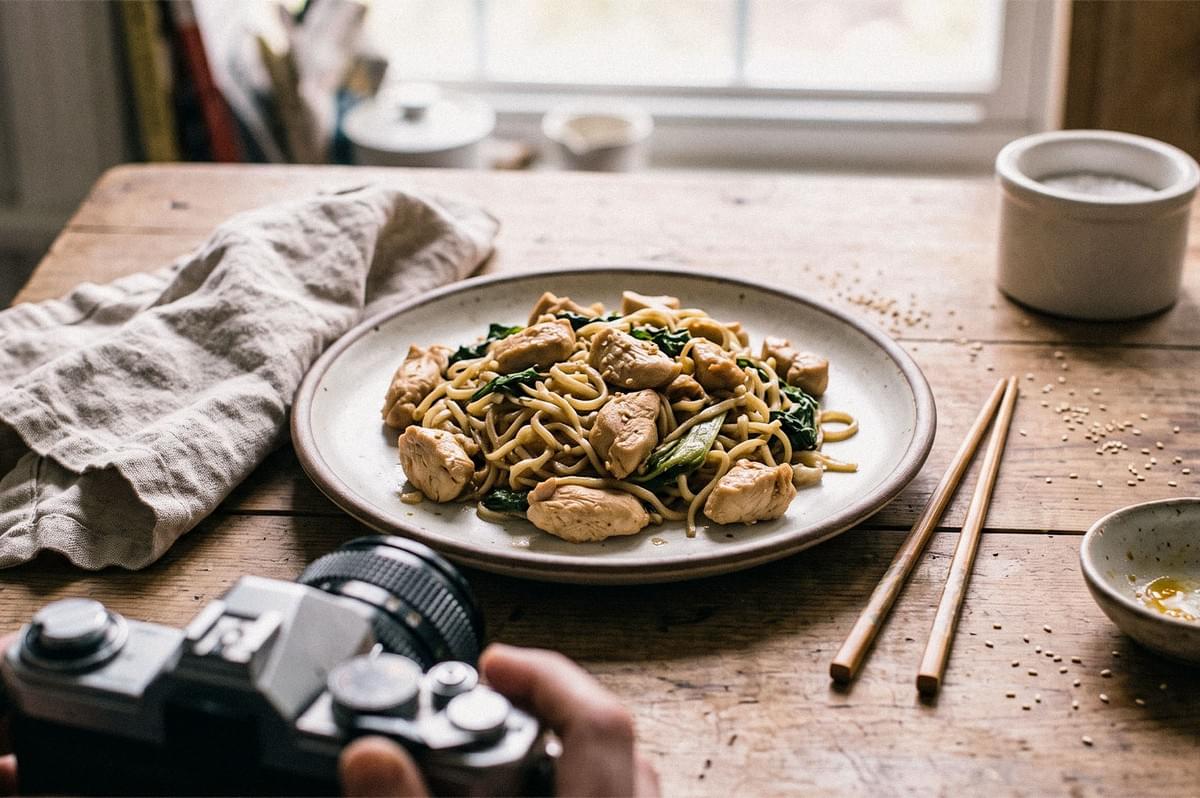 Plate of chicken noodles with leafy greens on a wooden table, photographed beside a camera, chopsticks, and a kitchen towel. Plate of chicken noodles with leafy greens on a wooden table, photographed beside a camera, chopsticks, and a kitchen towel.