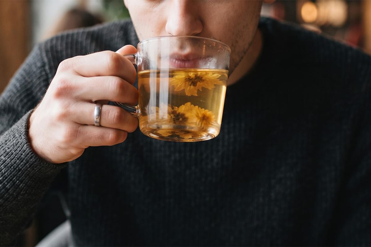 Man wearing a dark sweater holding and sipping a glass mug of hot chrysanthemum herbal tea. Man wearing a dark sweater holding and sipping a glass mug of hot chrysanthemum herbal tea.