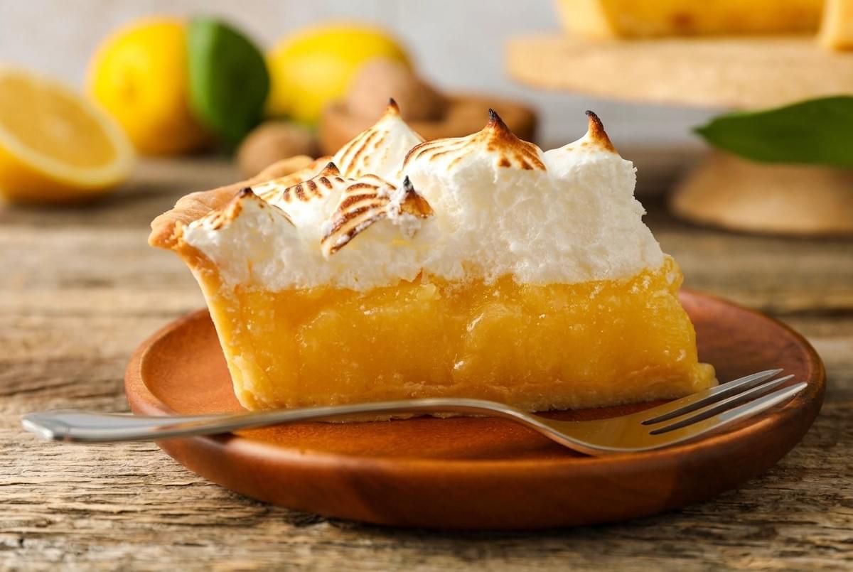 A slice of lemon meringue pie with toasted peaks served on a wooden plate with a fork. A slice of lemon meringue pie with toasted peaks served on a wooden plate with a fork.