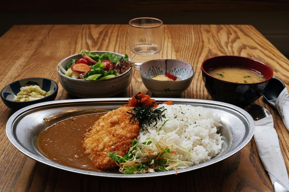 A meal featuring Japanese katsu curry served on a metal platter with white rice, dynamic shredded cabbage, and rich curry sauce. Surrounding side dishes include a fresh garden salad, miso soup, pickled vegetables, and a glass of water on a wooden table. A meal featuring Japanese katsu curry served on a metal platter with white rice, dynamic shredded cabbage, and rich curry sauce. Surrounding side dishes include a fresh garden salad, miso soup, pickled vegetables, and a glass of water on a wooden table.
