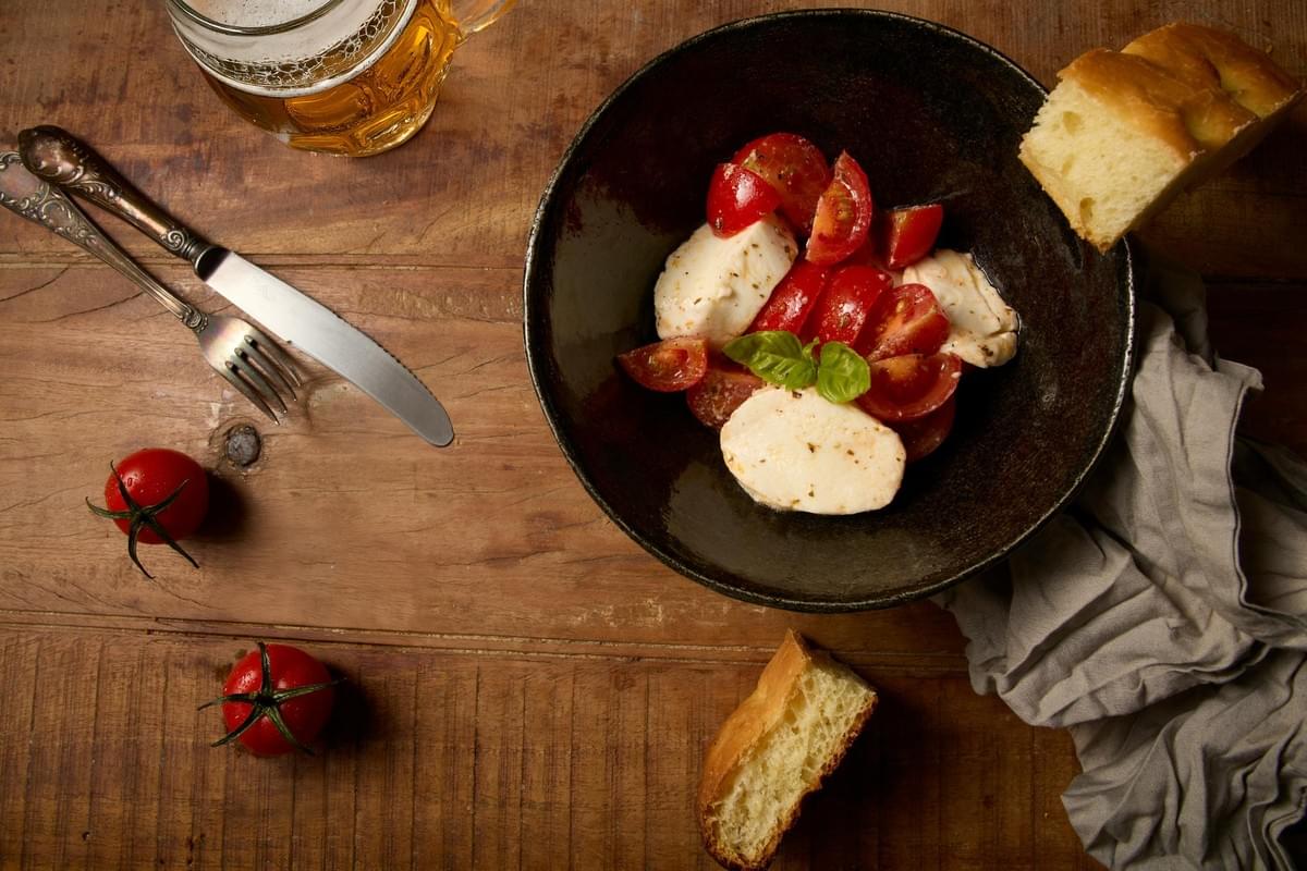 A top-down shot features a dark ceramic bowl filled with a Caprese salad of fresh mozzarella slices, juicy cherry tomatoes, and a single basil leaf. The bowl sits on a rustic wooden table alongside a glass of beer, a knife and fork, pieces of crusty bread, and loose tomatoes. A top-down shot features a dark ceramic bowl filled with a Caprese salad of fresh mozzarella slices, juicy cherry tomatoes, and a single basil leaf. The bowl sits on a rustic wooden table alongside a glass of beer, a knife and fork, pieces of crusty bread, and loose tomatoes.