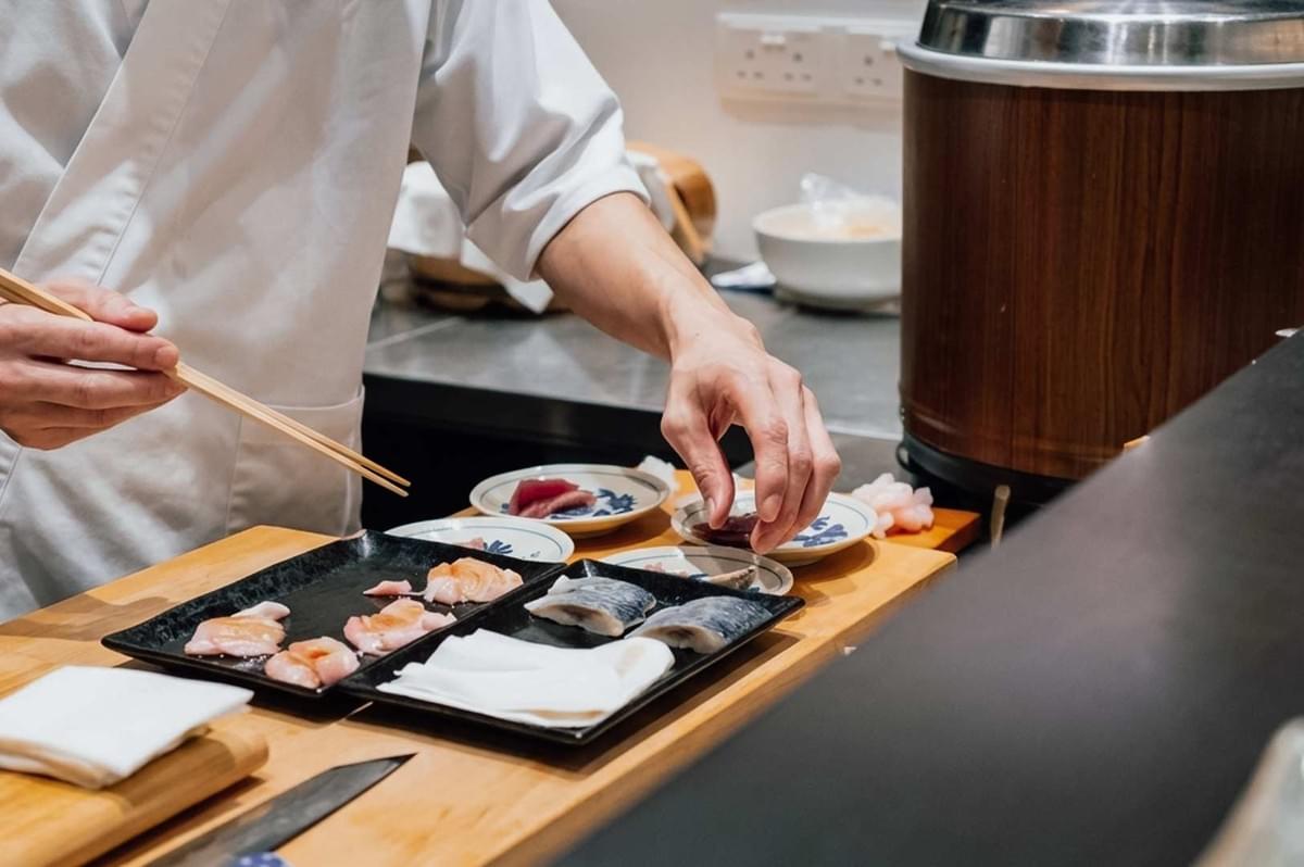A chef in a white uniform using chopsticks and precision hand techniques to meticulously arrange fresh fish slices—including salmon, mackerel, and tuna—onto small plates and dark rectangular platters along a wooden prep station. A chef in a white uniform using chopsticks and precision hand techniques to meticulously arrange fresh fish slices—including salmon, mackerel, and tuna—onto small plates and dark rectangular platters along a wooden prep station.