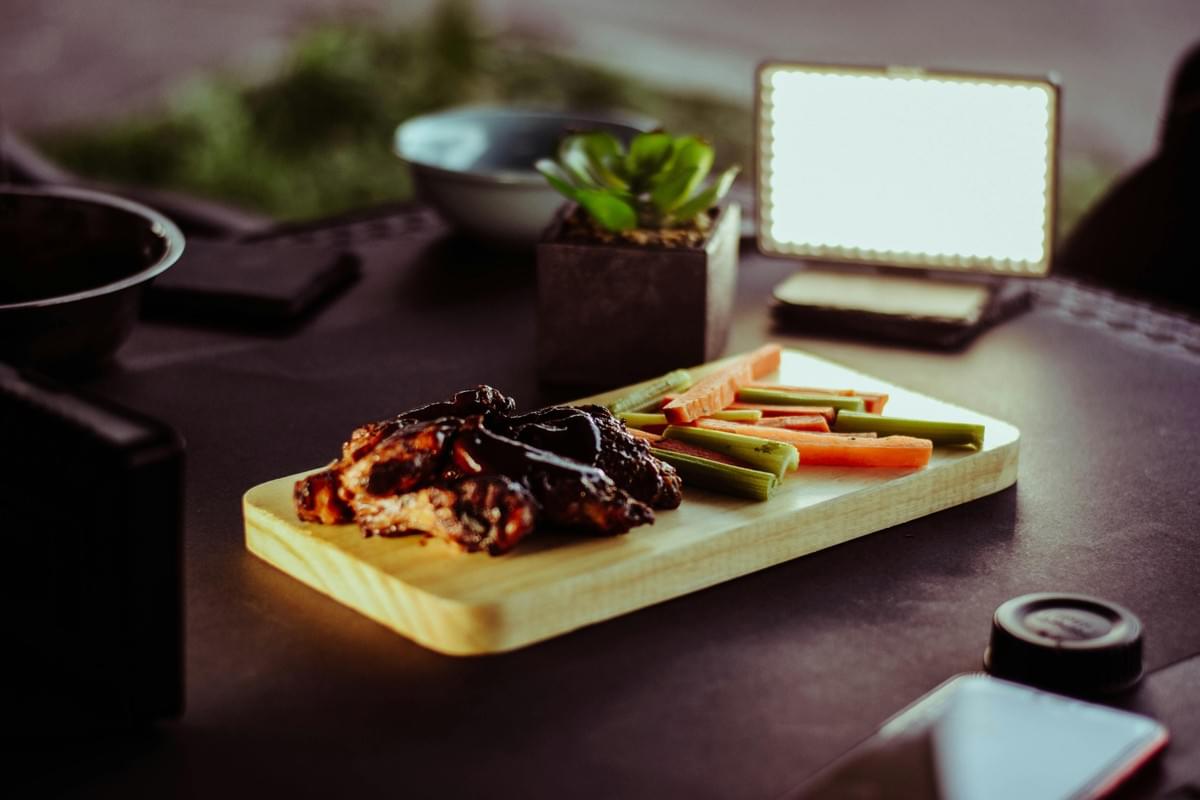A wooden serving board on a dark table holds a pile of dark, saucy chicken wings alongside sliced carrot and celery sticks. In the blurred background, a small potted succulent and a bright rectangular LED panel light illuminate the scene. A wooden serving board on a dark table holds a pile of dark, saucy chicken wings alongside sliced carrot and celery sticks. In the blurred background, a small potted succulent and a bright rectangular LED panel light illuminate the scene.