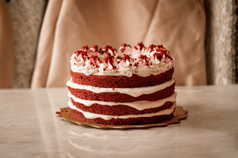 A whole four-layer red velvet naked cake showing exposed edges that display the cake's dense texture and rich color, topped with piped frosting and red cake crumbs. A whole four-layer red velvet naked cake showing exposed edges that display the cake's dense texture and rich color, topped with piped frosting and red cake crumbs.