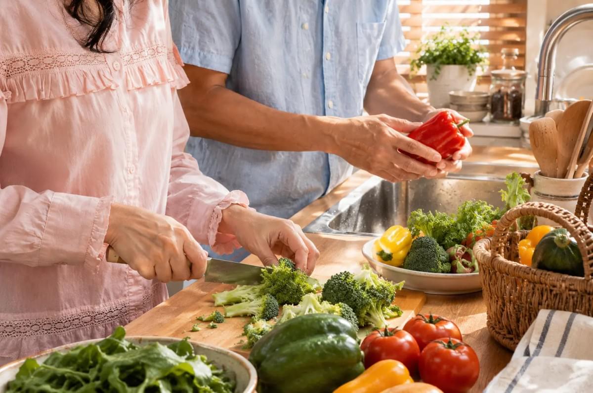 Two people chopping broccoli and prepping fresh vegetables together at a sunlit wooden kitchen counter. Two people chopping broccoli and prepping fresh vegetables together at a sunlit wooden kitchen counter.