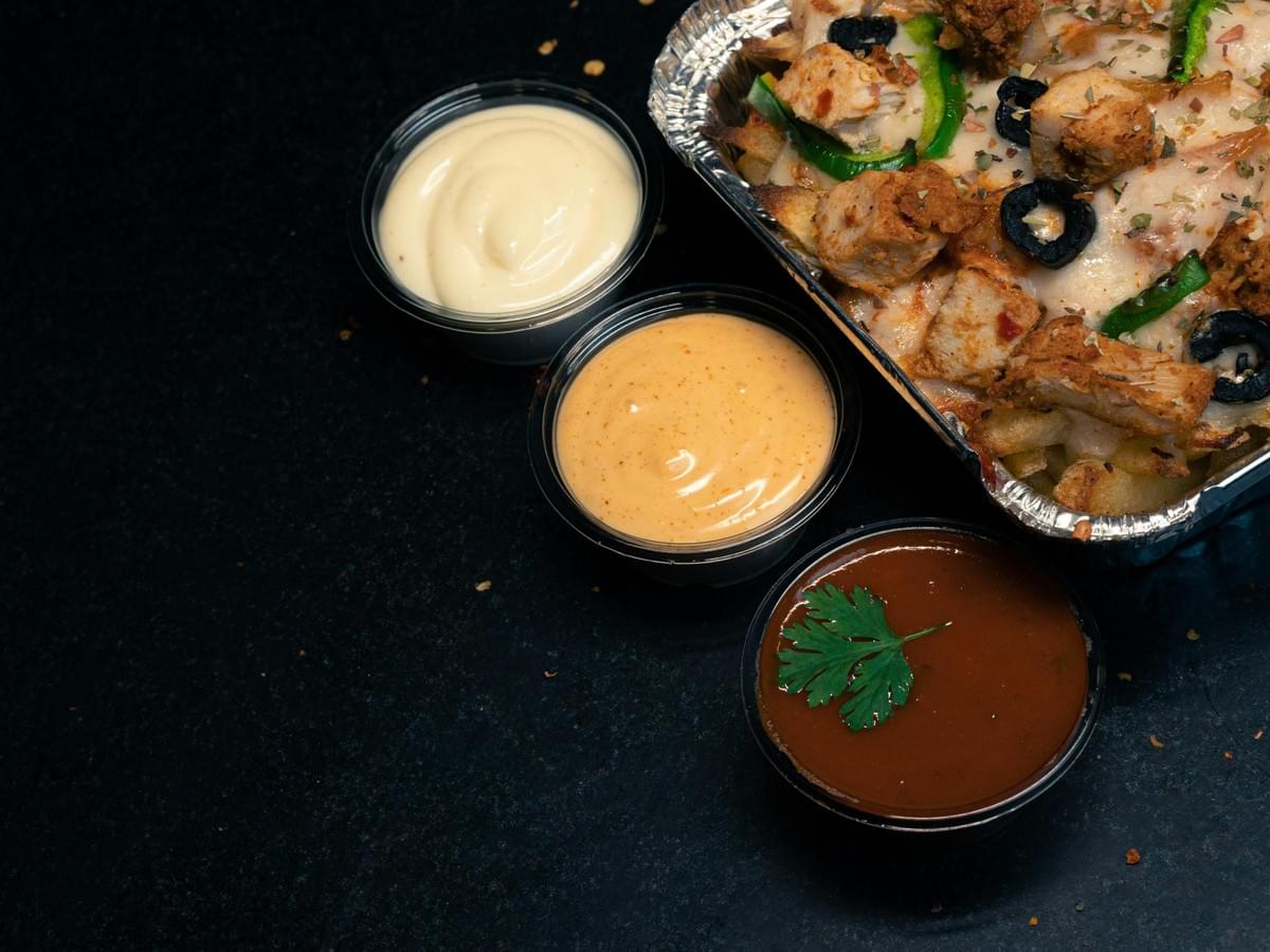 A aluminum container holds baked fries or nachos topped with melted cheese, seasoned chicken chunks, sliced green bell peppers, and black olives. Next to the dish, three small plastic cups contain garlic mayonnaise, a creamy orange sauce, and a brown sauce garnished with a fresh cilantro sprig. A aluminum container holds baked fries or nachos topped with melted cheese, seasoned chicken chunks, sliced green bell peppers, and black olives. Next to the dish, three small plastic cups contain garlic mayonnaise, a creamy orange sauce, and a brown sauce garnished with a fresh cilantro sprig.