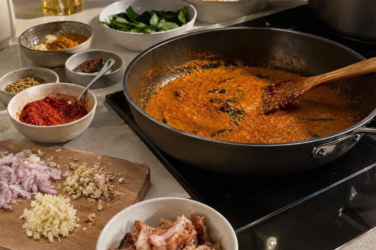 A cooking-in-progress shot shows a large skillet simmering with rich, orange laksa curry sauce on an induction cooktop, stirred by a wooden spatula. Surrounding the pan are prep bowls of spices, chili paste, leaves, and a wooden cutting board with chopped shallots, minced garlic, and sliced lemongrass. A cooking-in-progress shot shows a large skillet simmering with rich, orange laksa curry sauce on an induction cooktop, stirred by a wooden spatula. Surrounding the pan are prep bowls of spices, chili paste, leaves, and a wooden cutting board with chopped shallots, minced garlic, and sliced lemongrass.