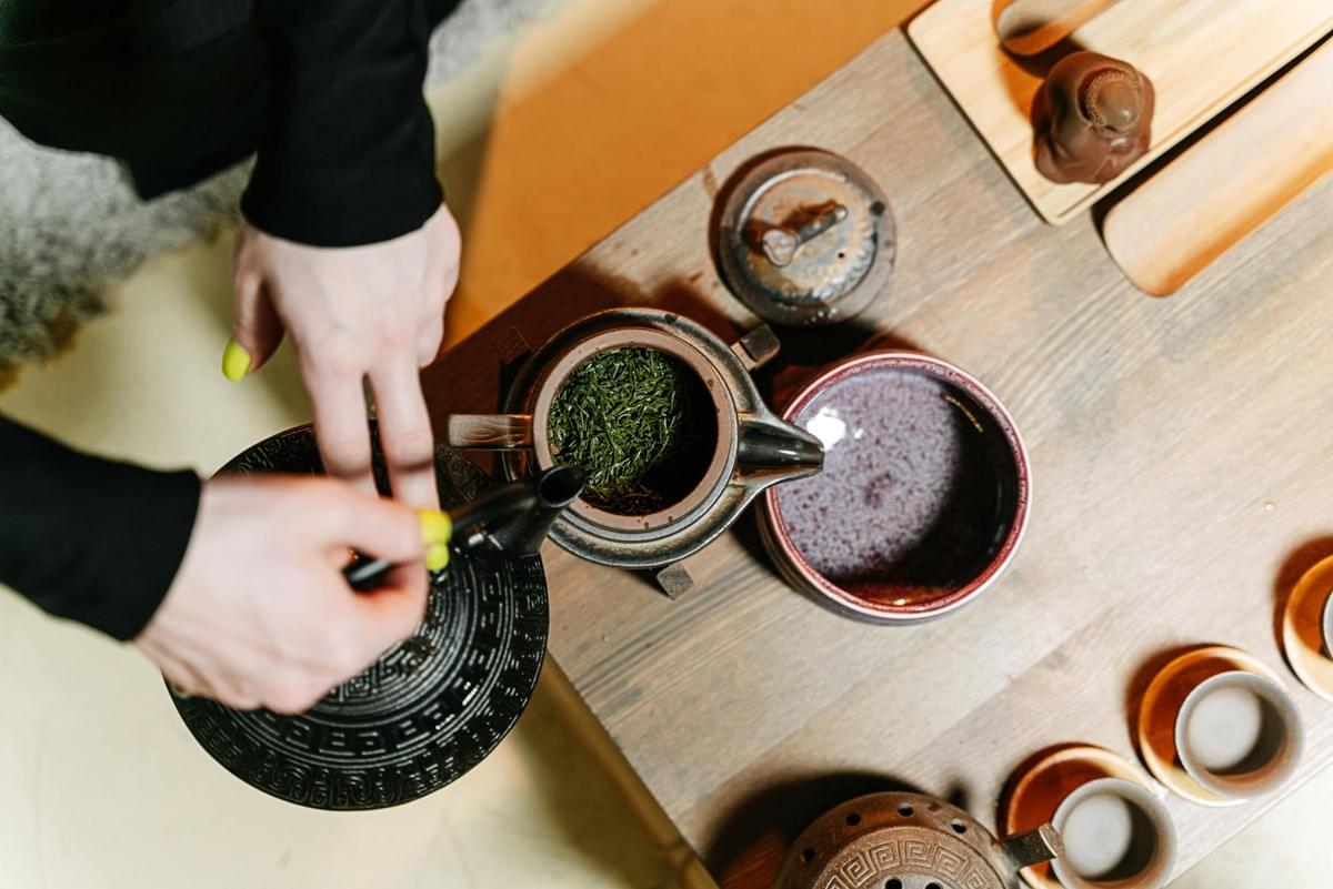 A vendor in a green shirt leans over tCaptured from an overhead angle, a person pours water from a textured black kettle into a teapot filled with vibrant loose green tea leaves. The wooden table is arranged for a traditional tea ceremony, featuring small ceramic cups, a serving pitcher, and various clay accessories.he counter of a brightly lit food stall to hand a meal to a customer in exchange for cash. The busy stand is plastered with colorful signs in Spanish, advertising fast food options like hot dogs, sandwiches, and fries. A vendor in a green shirt leans over tCaptured from an overhead angle, a person pours water from a textured black kettle into a teapot filled with vibrant loose green tea leaves. The wooden table is arranged for a traditional tea ceremony, featuring small ceramic cups, a serving pitcher, and various clay accessories.he counter of a brightly lit food stall to hand a meal to a customer in exchange for cash. The busy stand is plastered with colorful signs in Spanish, advertising fast food options like hot dogs, sandwiches, and fries.