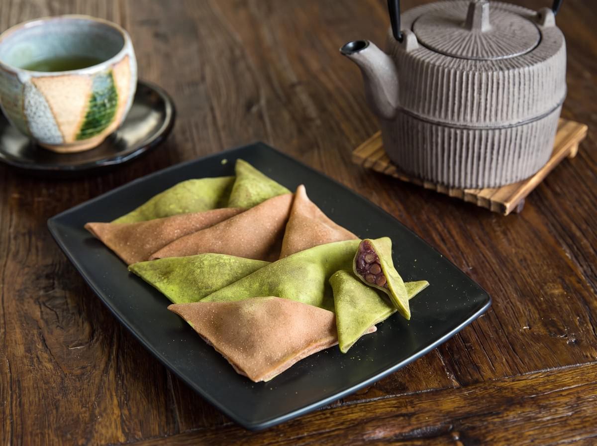 Assorted yatsuhashi mochi filled with red bean paste served on a black rectangular platter next to green tea. Assorted yatsuhashi mochi filled with red bean paste served on a black rectangular platter next to green tea.