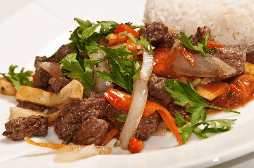 A side-angle shot of Lomo Saltado showing the glistening sauce on the beef and vegetables. The stir-fry is piled high next to a portion of white rice and heavily garnished with fresh flat-leaf parsley. A side-angle shot of Lomo Saltado showing the glistening sauce on the beef and vegetables. The stir-fry is piled high next to a portion of white rice and heavily garnished with fresh flat-leaf parsley.