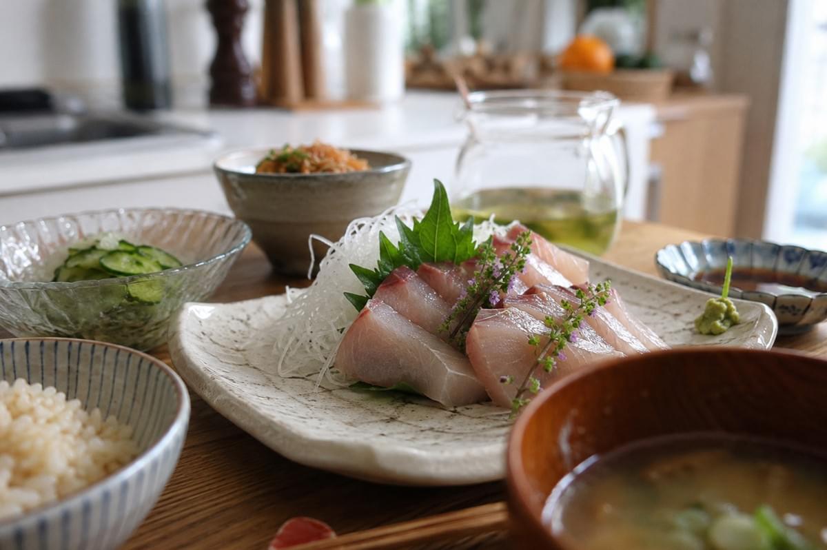 A beautifully plated serving of sashimi rests on a rustic, off-white ceramic dish, featuring thick slices of white fish atop a bed of delicate daikon radish threads and a shiso leaf. The fish is garnished with a small, flowering purple herb, and served alongside small bowls of rice, a bowl of soup, sliced cucumbers, and dipping sauces. The scene is captured in a homey, well-lit kitchen, highlighting a complete and balanced Japanese-style meal. A beautifully plated serving of sashimi rests on a rustic, off-white ceramic dish, featuring thick slices of white fish atop a bed of delicate daikon radish threads and a shiso leaf. The fish is garnished with a small, flowering purple herb, and served alongside small bowls of rice, a bowl of soup, sliced cucumbers, and dipping sauces. The scene is captured in a homey, well-lit kitchen, highlighting a complete and balanced Japanese-style meal.