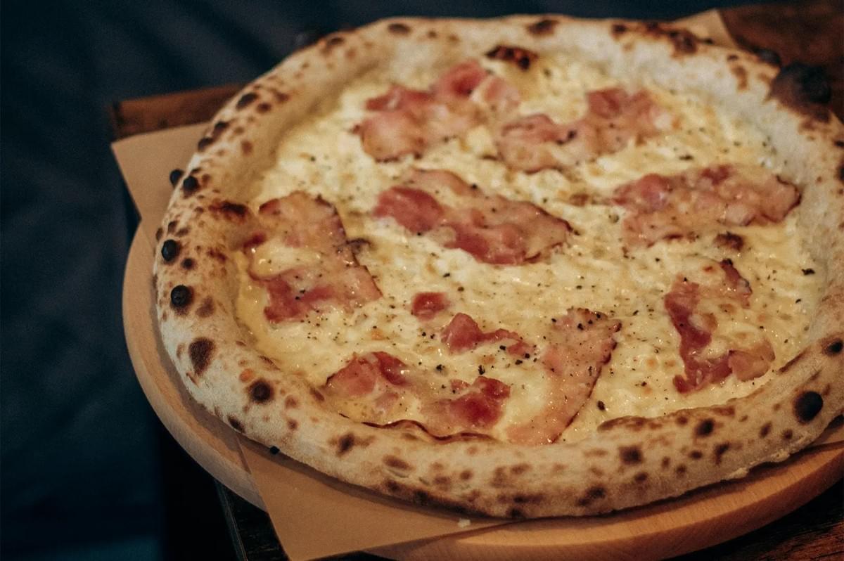 White base wood-fired pizza topped with crispy bacon and melted cheese on a wooden serving board. White base wood-fired pizza topped with crispy bacon and melted cheese on a wooden serving board.