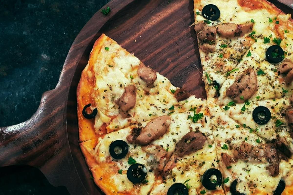A top-down shot shows a pizza with a missing slice on a wooden serving board. The pizza is topped with melted cheese, pieces of chicken, sliced black olives, and dried herbs. A top-down shot shows a pizza with a missing slice on a wooden serving board. The pizza is topped with melted cheese, pieces of chicken, sliced black olives, and dried herbs.