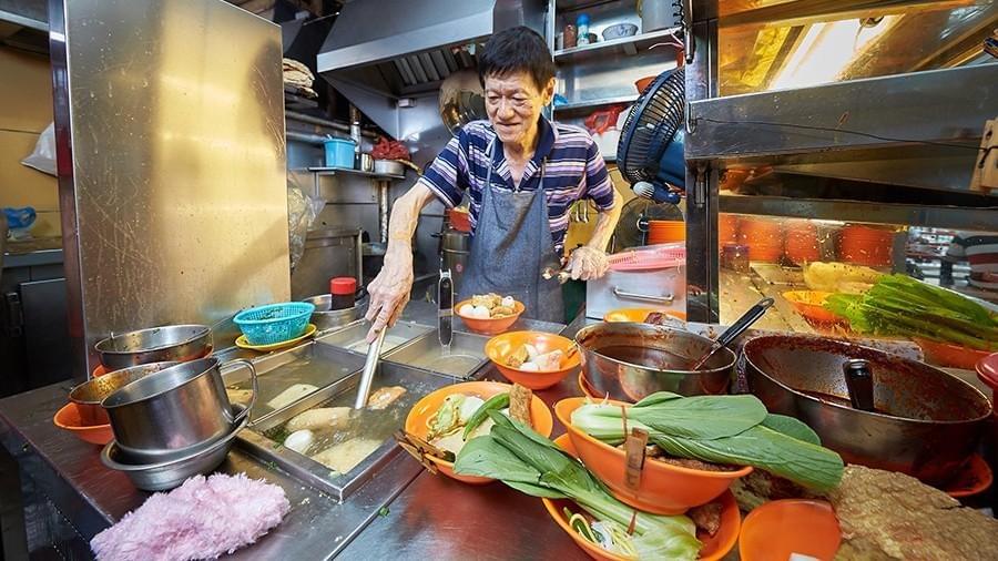 This image shows an elderly hawker meticulously preparing bowls of food at a bustling Singaporean food stall, surrounded by fresh ingredients and large pots of simmering broth. This image shows an elderly hawker meticulously preparing bowls of food at a bustling Singaporean food stall, surrounded by fresh ingredients and large pots of simmering broth.