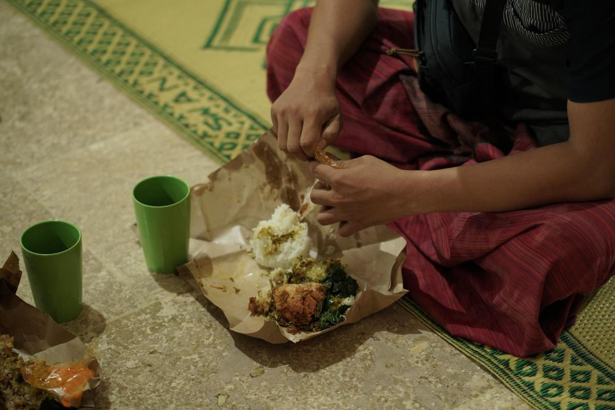 A person wearing red pants sits cross-legged on a patterned mat while preparing to eat a meal served on paper wrapping. Two green plastic cups sit nearby on the tiled floor next to the food portion of rice and dishes. A person wearing red pants sits cross-legged on a patterned mat while preparing to eat a meal served on paper wrapping. Two green plastic cups sit nearby on the tiled floor next to the food portion of rice and dishes.
