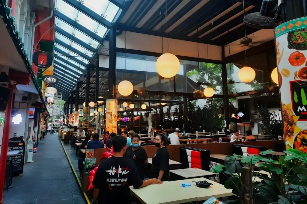 An outdoor, covered dining alley features a vibrant restaurant with large, glowing globe pendant lights and diners seated at tables. Servers in black shirts stand in the foreground, while a long, modern glass awning extends over the walkway to the left. An outdoor, covered dining alley features a vibrant restaurant with large, glowing globe pendant lights and diners seated at tables. Servers in black shirts stand in the foreground, while a long, modern glass awning extends over the walkway to the left.