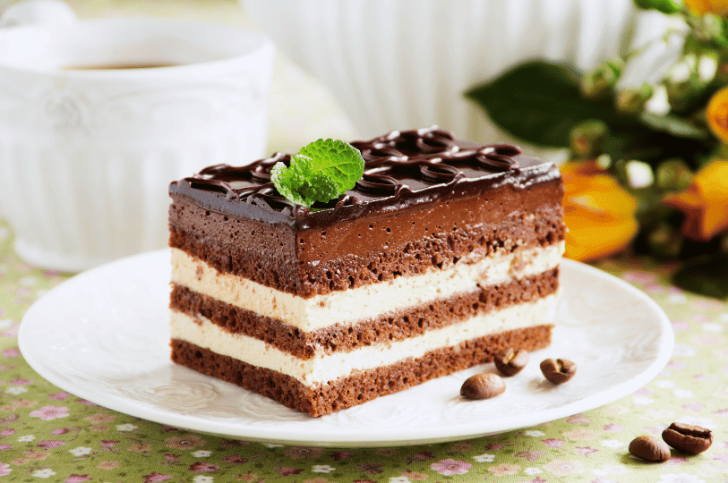 A thick slice of opera cake on a white plate, topped with decorative chocolate piping and a fresh mint sprig, with whole coffee beans scattered nearby on a floral tablecloth. A thick slice of opera cake on a white plate, topped with decorative chocolate piping and a fresh mint sprig, with whole coffee beans scattered nearby on a floral tablecloth.