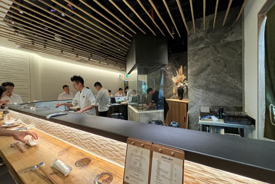 Chefs in white uniforms work behind an illuminated counter inside a modern Japanese restaurant with decorative wooden ceiling slats. Customers sit along the counter with menus and dining utensils placed in front of them. Chefs in white uniforms work behind an illuminated counter inside a modern Japanese restaurant with decorative wooden ceiling slats. Customers sit along the counter with menus and dining utensils placed in front of them.