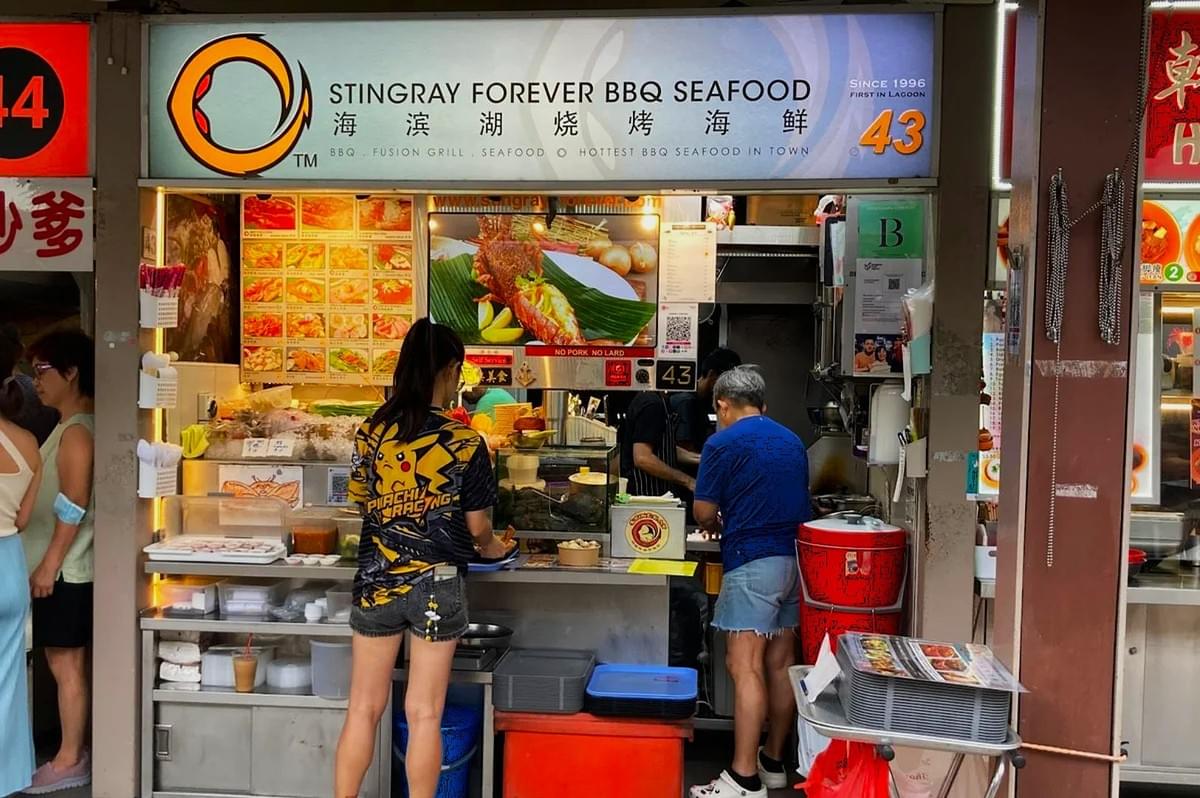 A bustling BBQ seafood stall named "Stingray Forever," displaying vibrant food photos. Two patrons stand at the counter, capturing a lively, casual atmosphere. A bustling BBQ seafood stall named "Stingray Forever," displaying vibrant food photos. Two patrons stand at the counter, capturing a lively, casual atmosphere.