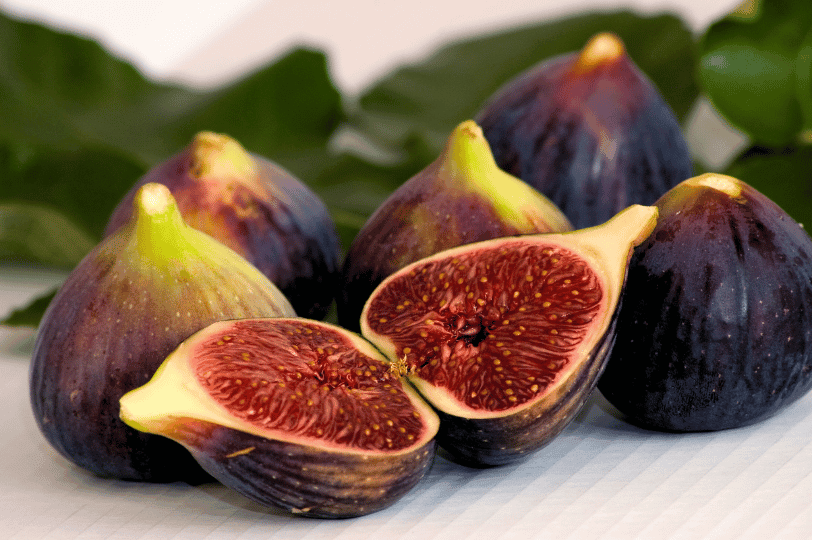 Two halves of a ripe mission fig displayed prominently in the foreground, surrounded by five whole figs and green leaves on a clean, white textured background. Two halves of a ripe mission fig displayed prominently in the foreground, surrounded by five whole figs and green leaves on a clean, white textured background.