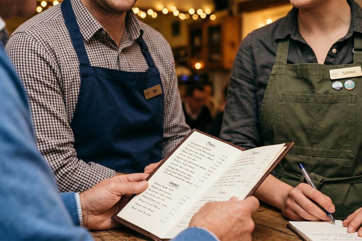Restaurant customer looking at a dinner menu while two waitstaff servers in aprons take the order. Restaurant customer looking at a dinner menu while two waitstaff servers in aprons take the order.