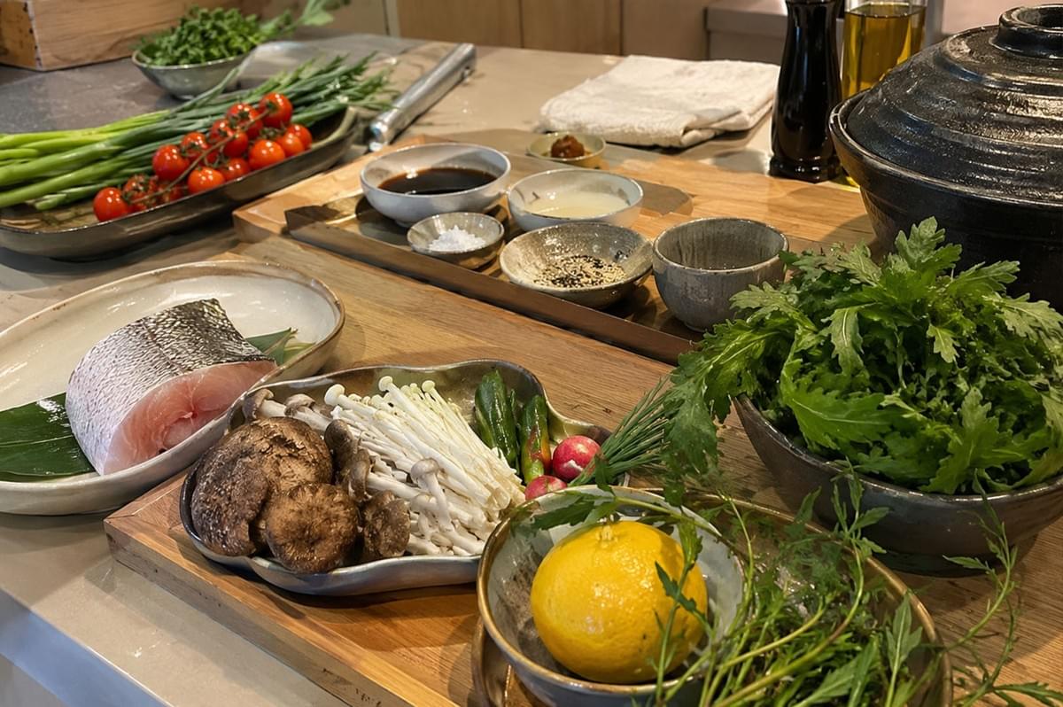 A wooden kitchen prep counter is filled with fresh cooking ingredients laid out in ceramic bowls and platters. In the foreground, a thick cut of fresh fish rests on a leaf inside an oval dish next to a bowl of assorted mushrooms including shiitake and enoki, a bowl containing a whole yellow citrus fruit with fresh herbs, and a large dish packed with vibrant green leafy vegetables. The background features scallions, cherry tomatoes on the vine, small pinch bowls of seasonings and sauces, and a large black ceramic donabe pot. A wooden kitchen prep counter is filled with fresh cooking ingredients laid out in ceramic bowls and platters. In the foreground, a thick cut of fresh fish rests on a leaf inside an oval dish next to a bowl of assorted mushrooms including shiitake and enoki, a bowl containing a whole yellow citrus fruit with fresh herbs, and a large dish packed with vibrant green leafy vegetables. The background features scallions, cherry tomatoes on the vine, small pinch bowls of seasonings and sauces, and a large black ceramic donabe pot.