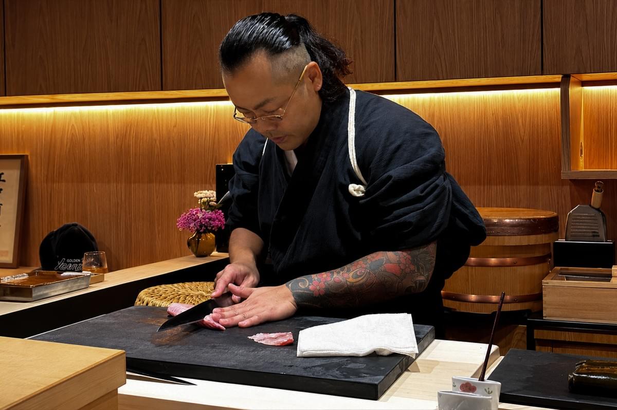 A medium close-up shot captures a focused sushi chef, with his dark hair tied back in a ponytail and shaved on the sides, leaning over a black cutting board to slice raw fish. He wears thin-rimmed glasses and a dark blue, textured wrap-style uniform with a white cord tied over his left shoulder, revealing a vibrant, multi-colored floral tattoo completely covering his left forearm. With precision, he uses a long, professional sushi knife held in his right hand to cut a piece of pink fish, stabilizing it with his left hand, while a clean white cloth rests nearby on the board. The warm-toned background features elegant wood paneling accented by soft under-cabinet lighting, a small vase of pink flowers, and traditional wooden sushi kitchen equipment like a rice tub to his right. A medium close-up shot captures a focused sushi chef, with his dark hair tied back in a ponytail and shaved on the sides, leaning over a black cutting board to slice raw fish. He wears thin-rimmed glasses and a dark blue, textured wrap-style uniform with a white cord tied over his left shoulder, revealing a vibrant, multi-colored floral tattoo completely covering his left forearm. With precision, he uses a long, professional sushi knife held in his right hand to cut a piece of pink fish, stabilizing it with his left hand, while a clean white cloth rests nearby on the board. The warm-toned background features elegant wood paneling accented by soft under-cabinet lighting, a small vase of pink flowers, and traditional wooden sushi kitchen equipment like a rice tub to his right.