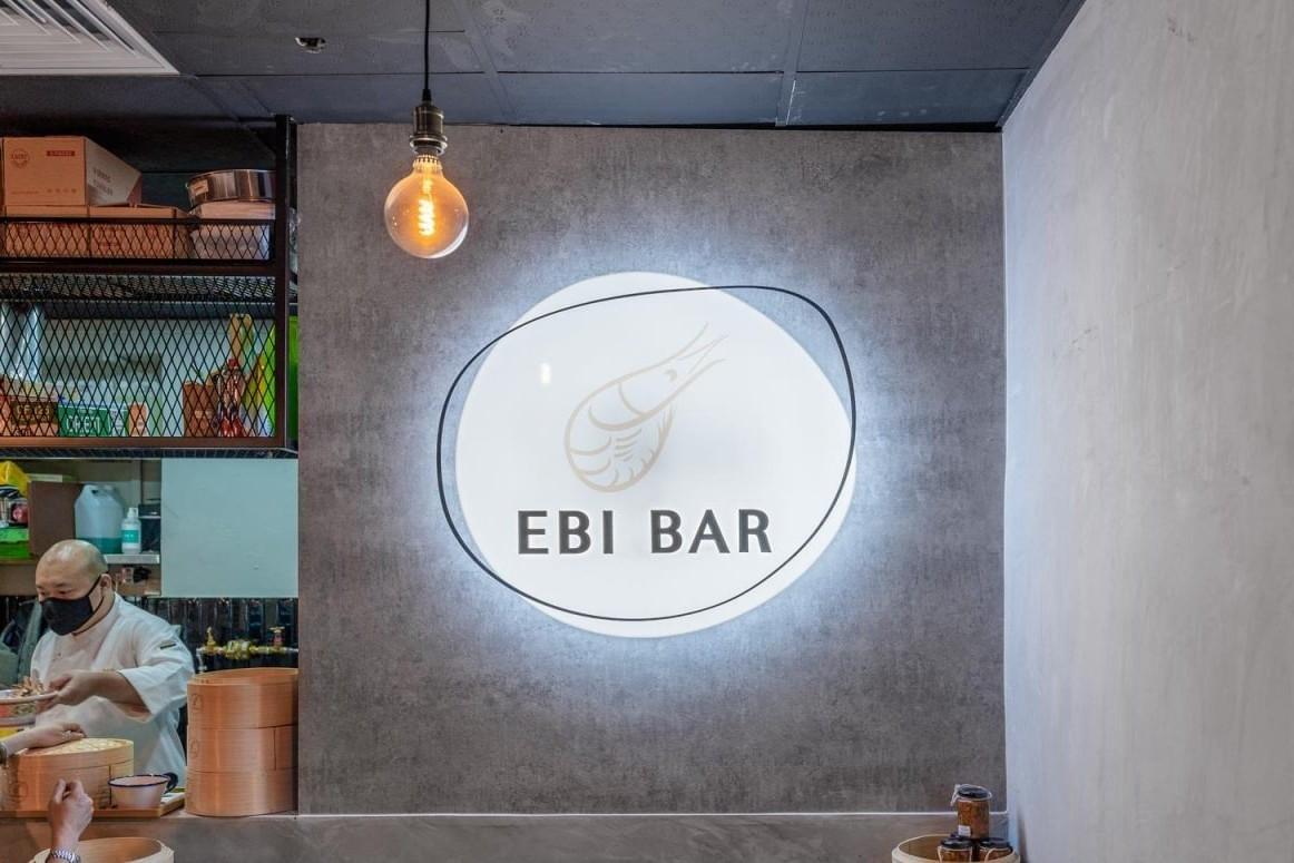 An illuminated circular sign displaying the name "EBI BAR" and a stylized shrimp illustration is mounted on a concrete wall. To the left, a chef in a face mask works behind the counter beneath a warm, hanging light bulb. An illuminated circular sign displaying the name "EBI BAR" and a stylized shrimp illustration is mounted on a concrete wall. To the left, a chef in a face mask works behind the counter beneath a warm, hanging light bulb.