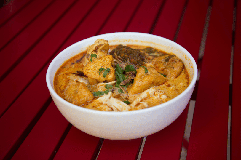 A white bowl of laksa sitting on a red slatted table, filled with a spicy coconut broth, chicken pieces, porous tofu puffs, and a spoonful of dark sambal paste. A white bowl of laksa sitting on a red slatted table, filled with a spicy coconut broth, chicken pieces, porous tofu puffs, and a spoonful of dark sambal paste.