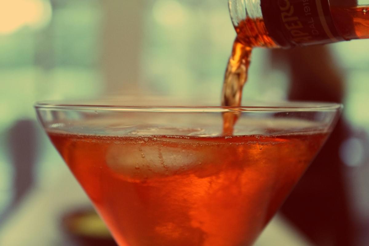 A close-up shot captures a vibrant orange liquid being poured from a bottle into a wide-rimmed cocktail glass filled with ice. The background is softly blurred, emphasizing the warm, effervescent texture of the drink. A close-up shot captures a vibrant orange liquid being poured from a bottle into a wide-rimmed cocktail glass filled with ice. The background is softly blurred, emphasizing the warm, effervescent texture of the drink.