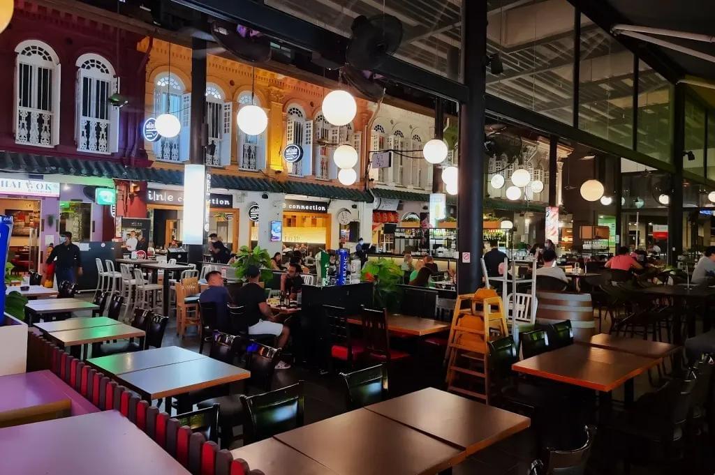 The image shows an open-air dining area at night, filled with numerous wooden tables, chairs, and patrons enjoying meals. Large, glowing white globe lights illuminate the seating area, with a vibrant row of colorful, historic shop-houses visible in the background. The image shows an open-air dining area at night, filled with numerous wooden tables, chairs, and patrons enjoying meals. Large, glowing white globe lights illuminate the seating area, with a vibrant row of colorful, historic shop-houses visible in the background.